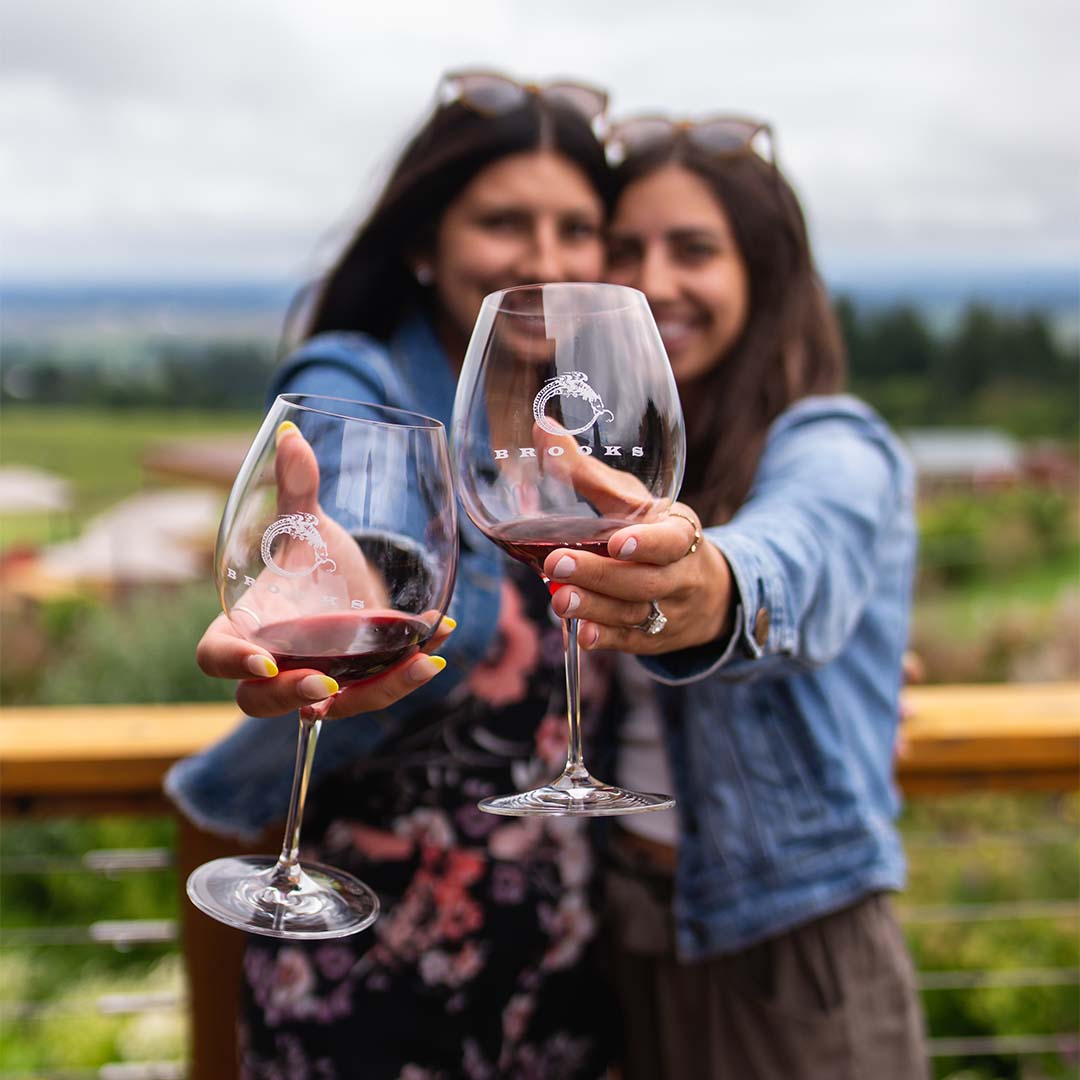 Two young women smiling and clinking glasses of Brooks Red Wine while on the deck overlooking mountains.