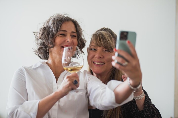 A smartly-dressed woman with gray curly hair holds up a glass of Riesling as she smiles for a selfie with a long-haired blonde woman. 