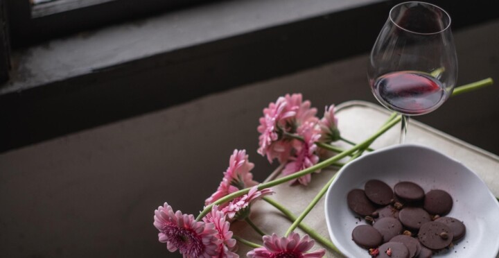 Pink flowers lay next to a bowl of dark chocolate disks and a glass of Pinot Noir as part of a Galentine's Day dinner course. 