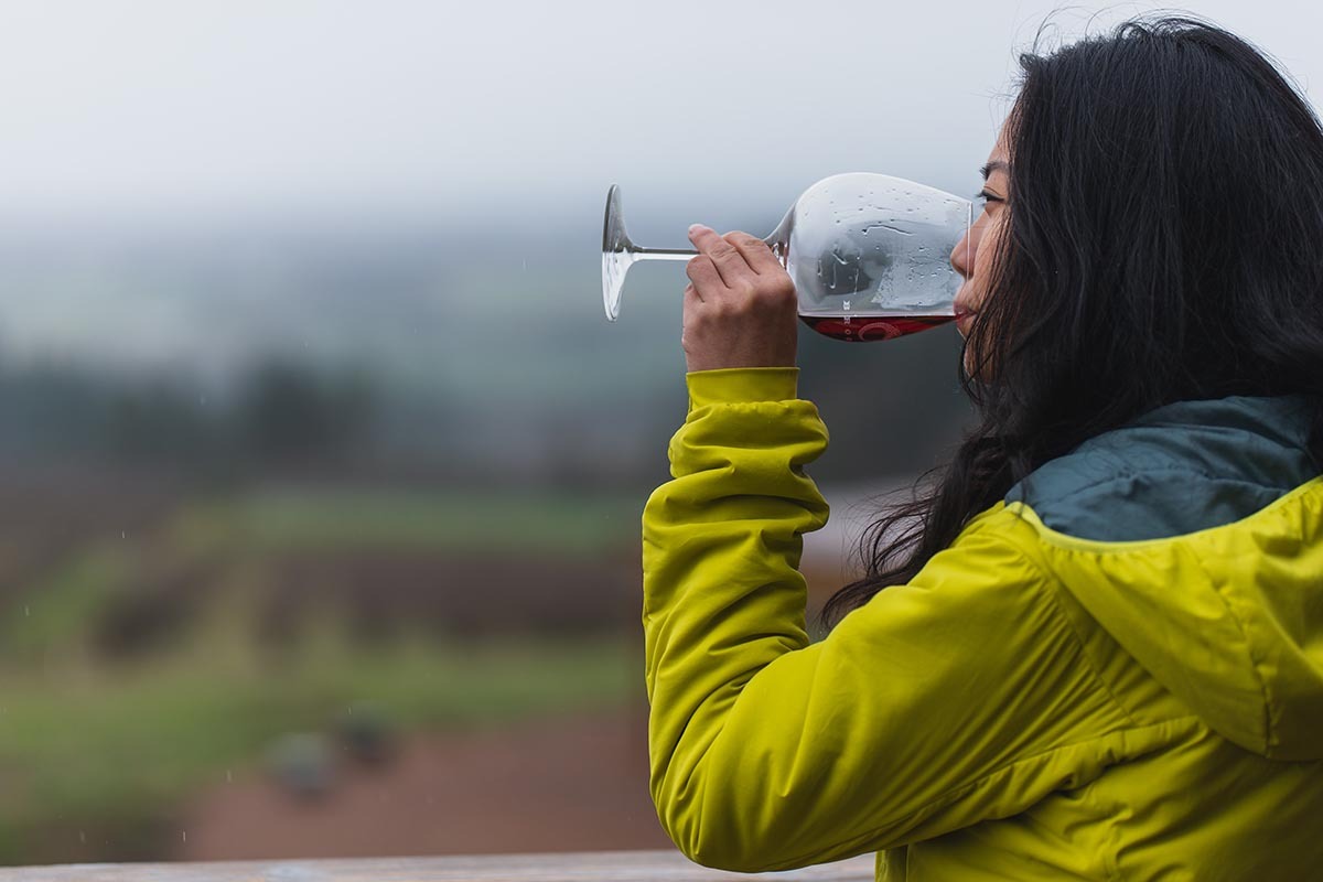 On a rainy day, a woman in a bright yellow raincoat takes a sip of Pinot Noir while looking out at Brooks Estate Vineyard, one of six top Oregon wineries. 
