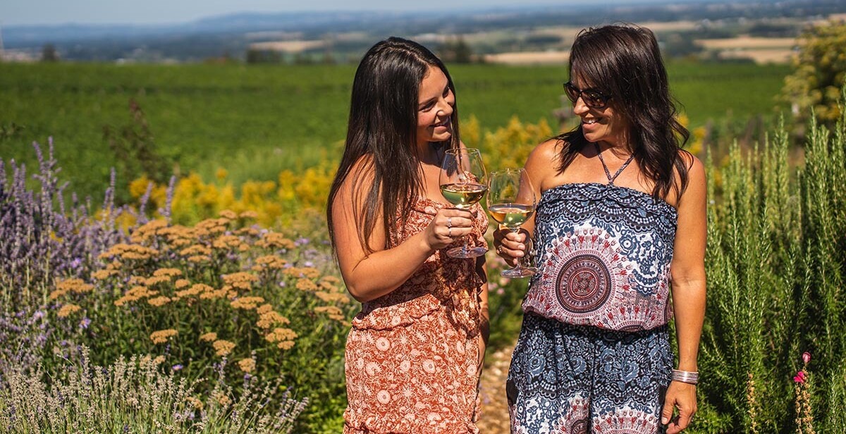 Two young women pose for an Instagram photo while tasting wine and walking through the lush garden at Brooks Wine on a sunny day.