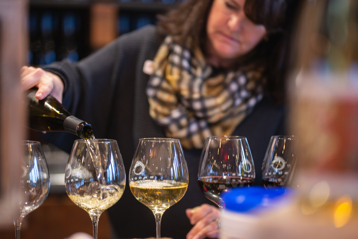 A brunette woman wearing a black sweater and yellow plaid scarf pours a flight of non-alcoholic wine for a blind tasting.