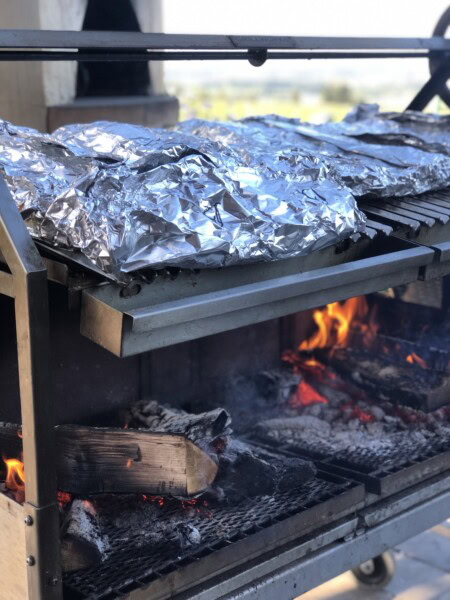 Foil packets of wild-caught salmon sit atop a grill over hot flaming logs of wood at a BBQ bash. 