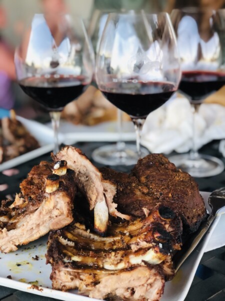 A plate of bbq grilled ribs sits in front of several glasses of red wine at a summer party.