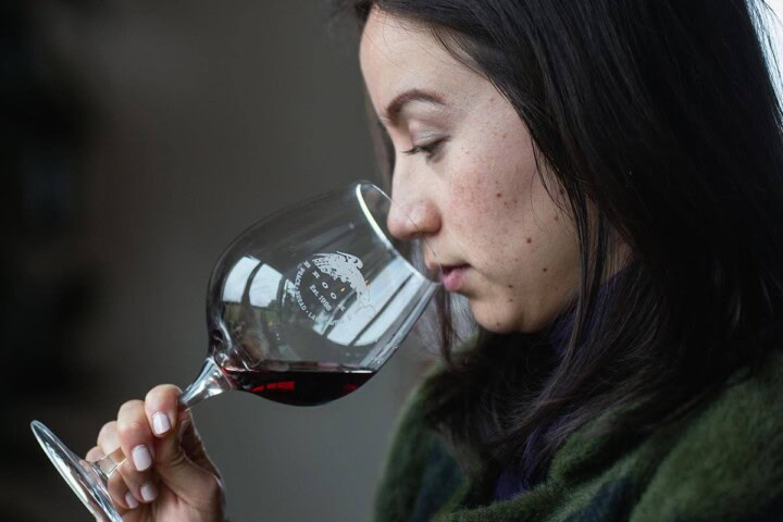 A young Asian woman with dark hair intently smells a glass of red wine as she determines how long to decant. 