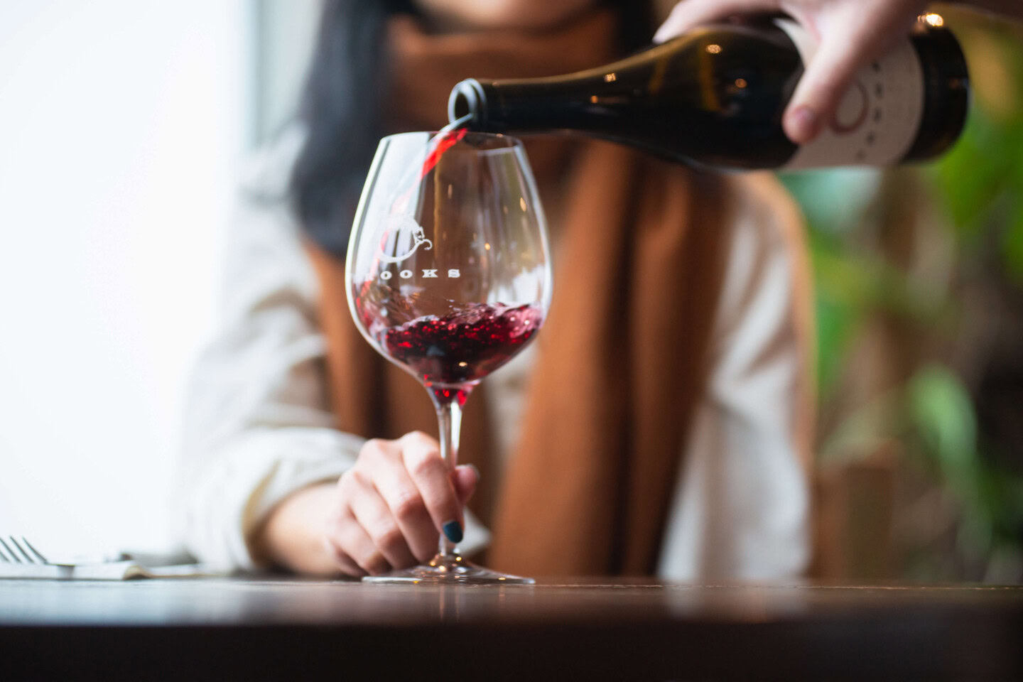 A dark-haired woman wearing a white blouse and brown scarf is poured a glass of Pinot Noir as she holds the glass by the stem in observance of wine etiquette.