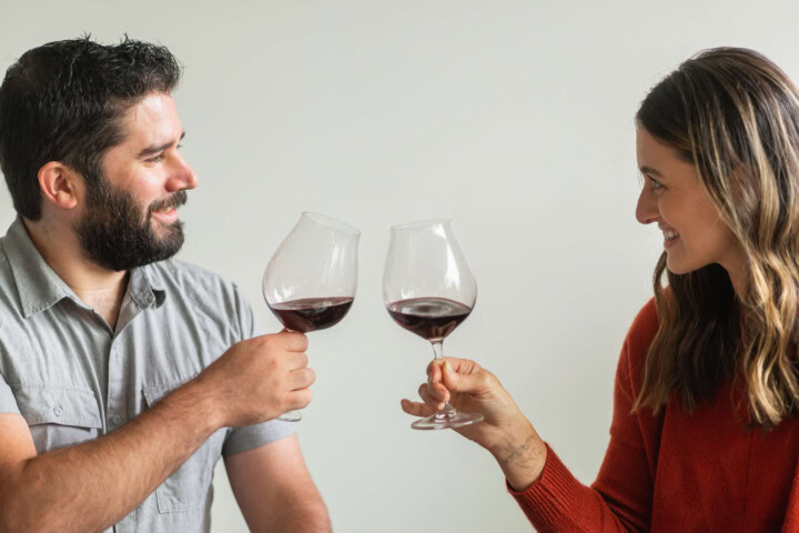 A dark-haired man in a gray shirt and a blonde woman in a red sweater smile at one another as they toast two glasses of Pinot Noir. 