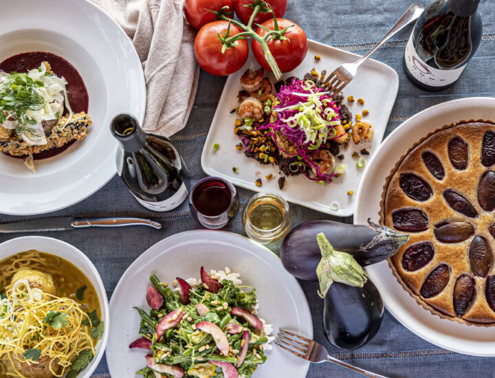 Light blue table cloth set with a wine and food pairing lunch including a plum tart, string bean salad with feta cheese,  corn and corn fritter, and crispy pork loin with shaved fennel.