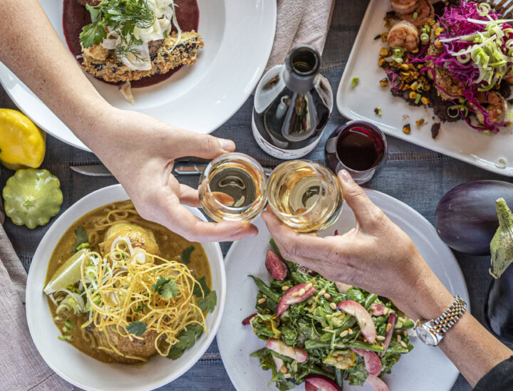 Two hands toast glasses of white wine above a table set with Thai green curry and a spring salad with string beans and white peaches. 