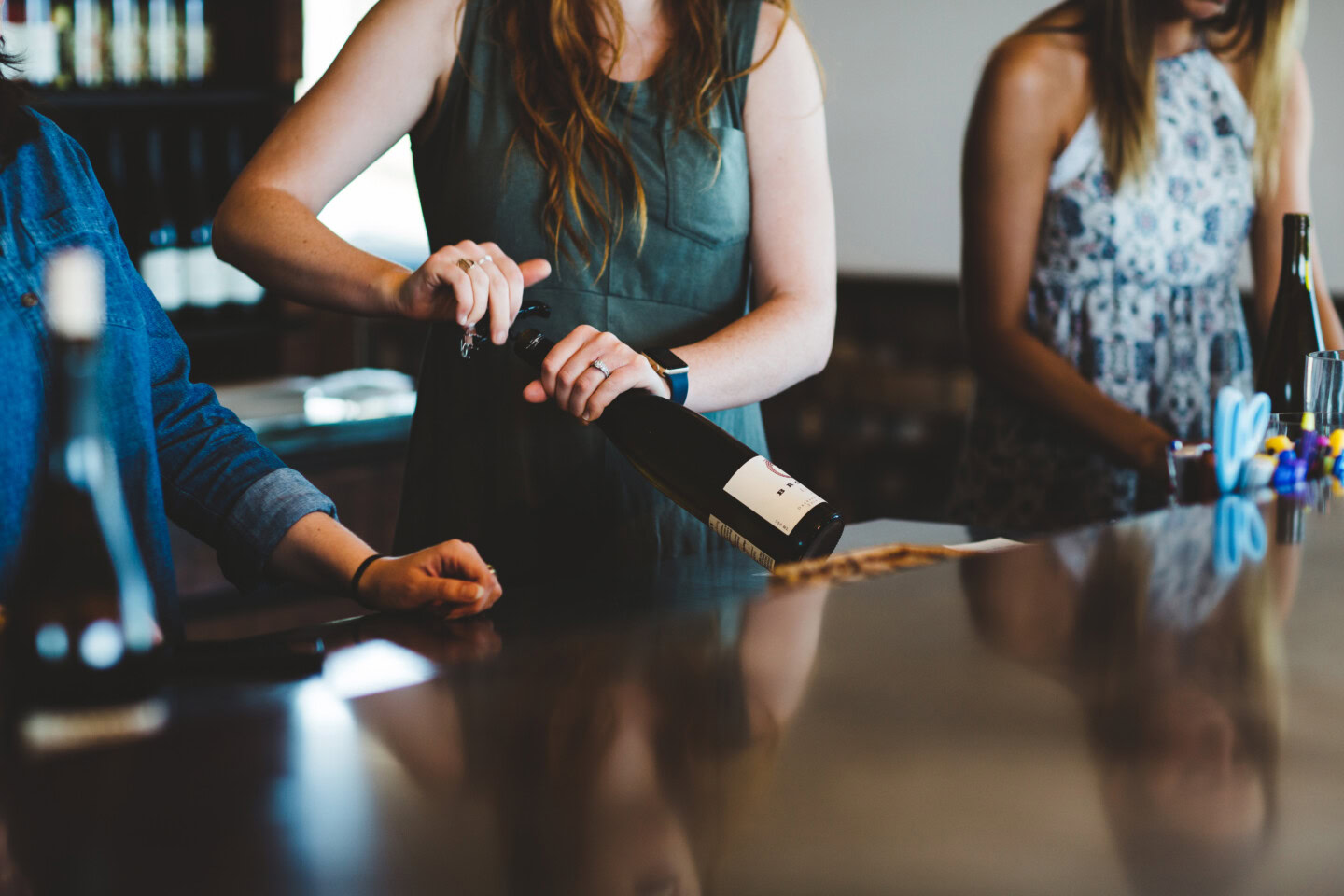 A series of women sommeliers standing at a winery tasting room bar are opening wine bottles for the day's wine flights. 