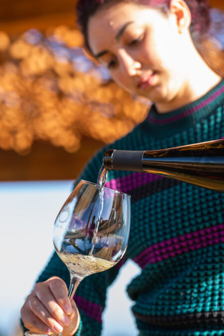 A young brunette woman in a dark blue sweater caringly pours a glass of Riesling from Brooks Wine. 